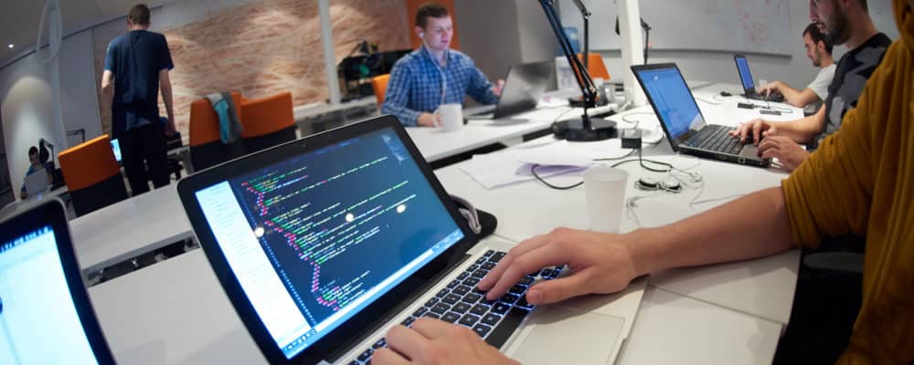 Bilden visar en bärbar dator på ett bord i ett kontor, och händer som jobbar på tangentbordet